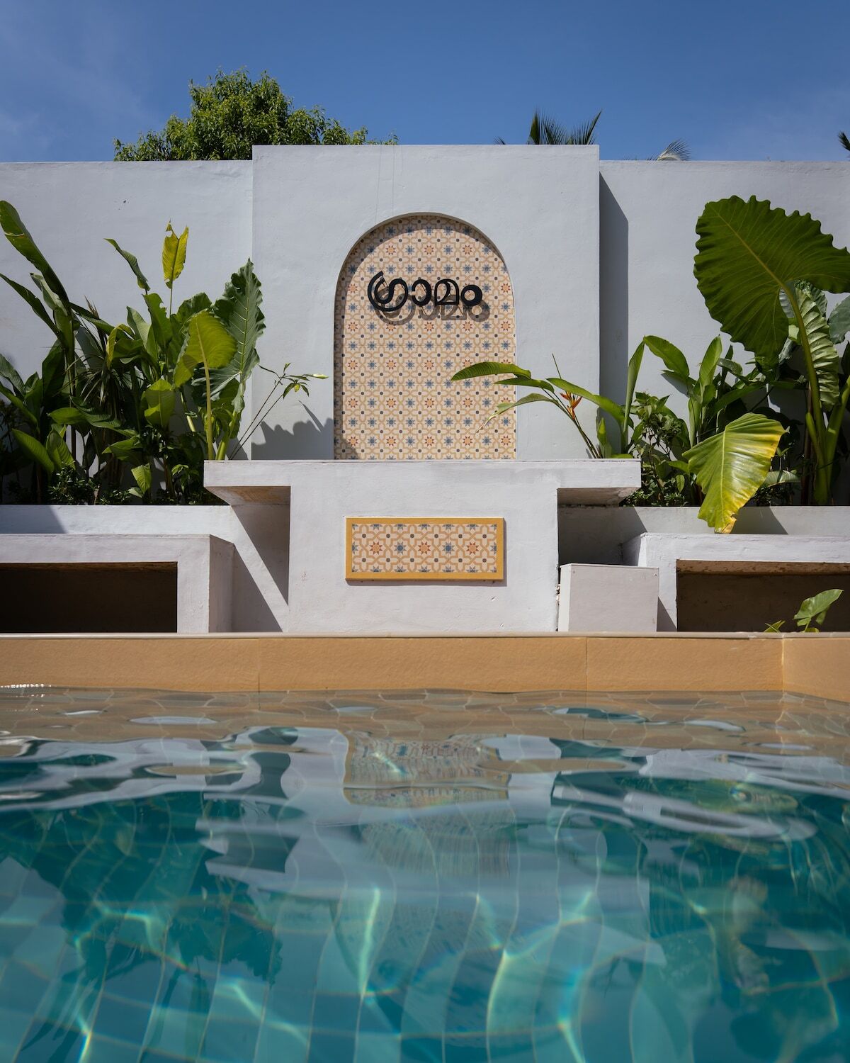 The villa's private pool in daylight, enclosed by a white lime-plaster wall with a tiled arch carrying the villa's name in Malayalam, framed by travellers' palms.